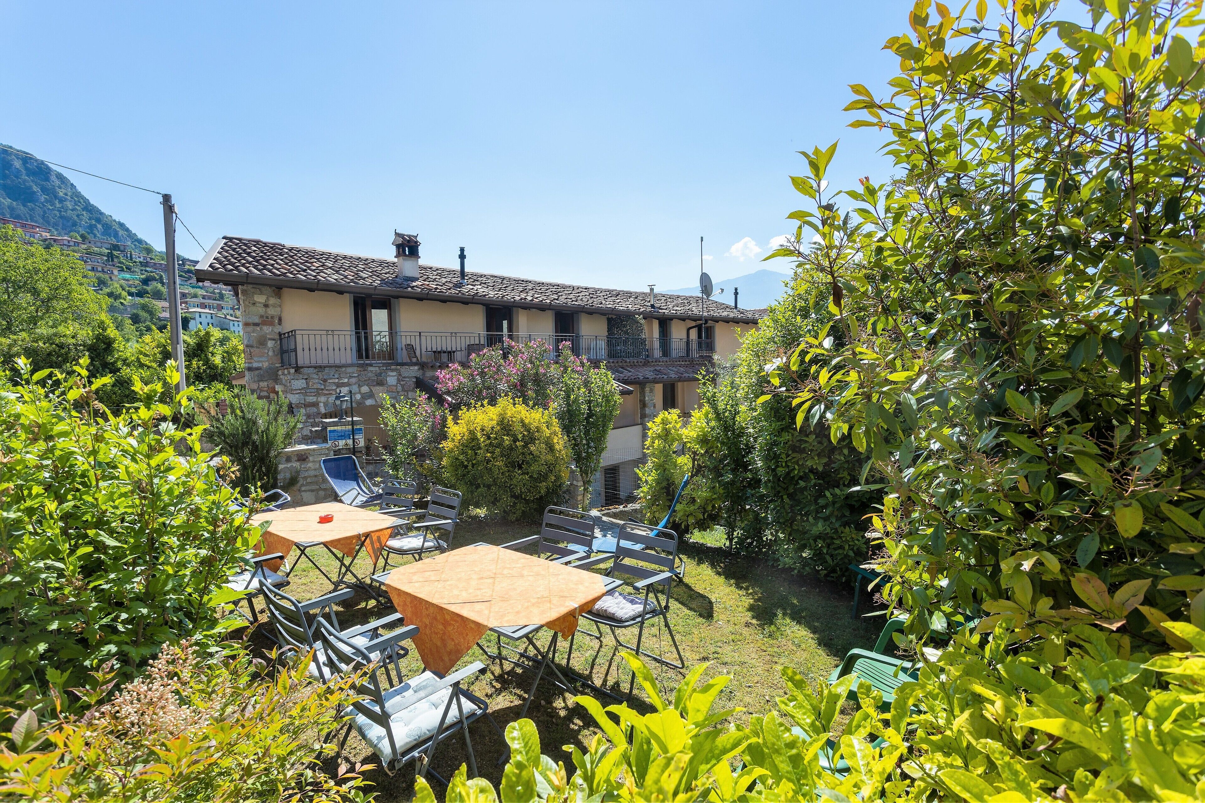 Une cour avec une table et des chaises, entourée de verdure et bordée d’un bâtiment dont le toit est recouvert de tuiles.