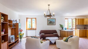 Ett vardagsrum med en soffa, fåtöljer, en tv, en bokhylla och en matplats, med ett kök synligt i bakgrunden.