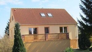 Une maison à deux étages, avec un toit recouvert de tuiles rouges, un balcon doté d’une balustrade en bois et une petite fenêtre à l’étage supérieur.