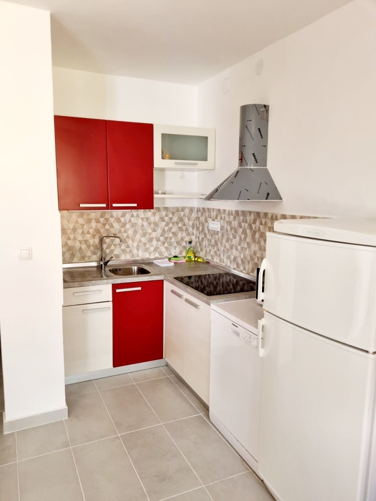 A compact kitchen with red cabinets, a built-in microwave, a stainless steel sink, and a white refrigerator.