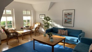 Een gezellige woonkamer met een blauwe bank, rieten stoelen, een houten salontafel en een groot raam met gordijnen.
