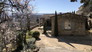 Un bâtiment en pierre avec un toit de tuiles, entouré d’un chemin de gravier et d’arbres en fleurs.