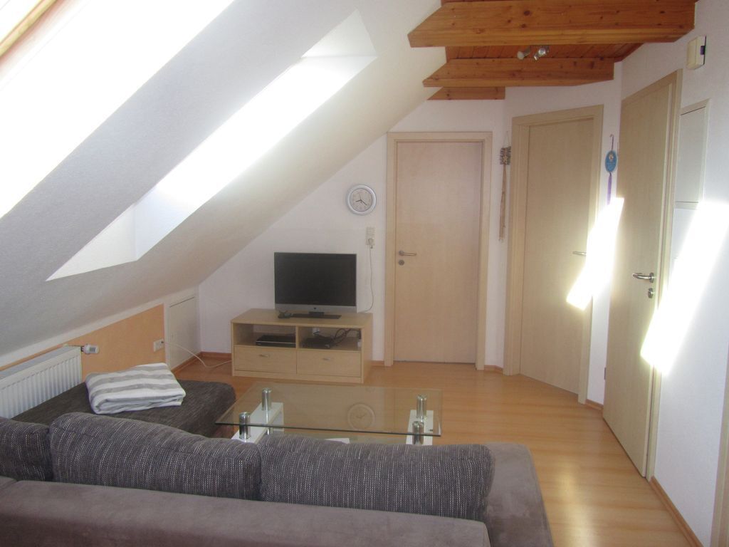 Ett rum i loftstil med ett sluttande tak, en platt-TV på ett trästativ, ett glasbord, en soffa och en dörr med en ljusströmbrytare.