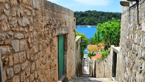 A narrow stone pathway leading to a building with a green door, surrounded by stone walls and a view of a body of water and greenery in the background. - Room in Cavtat with Seaview, Balcony, Air condition, WIFI (3686-1) (Cavtat)