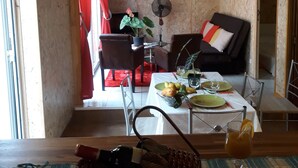 A cozy living room with a dining area, wooden walls, a red curtain, a fan, and a dining table set with plates and a bottle. - la cariole (Saint-Pierre)