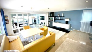 A modern living room with a yellow sofa, a dining table, and a kitchen area with a TV and shelves. - Luxurious holiday home directly on the lake (Wervershoof)