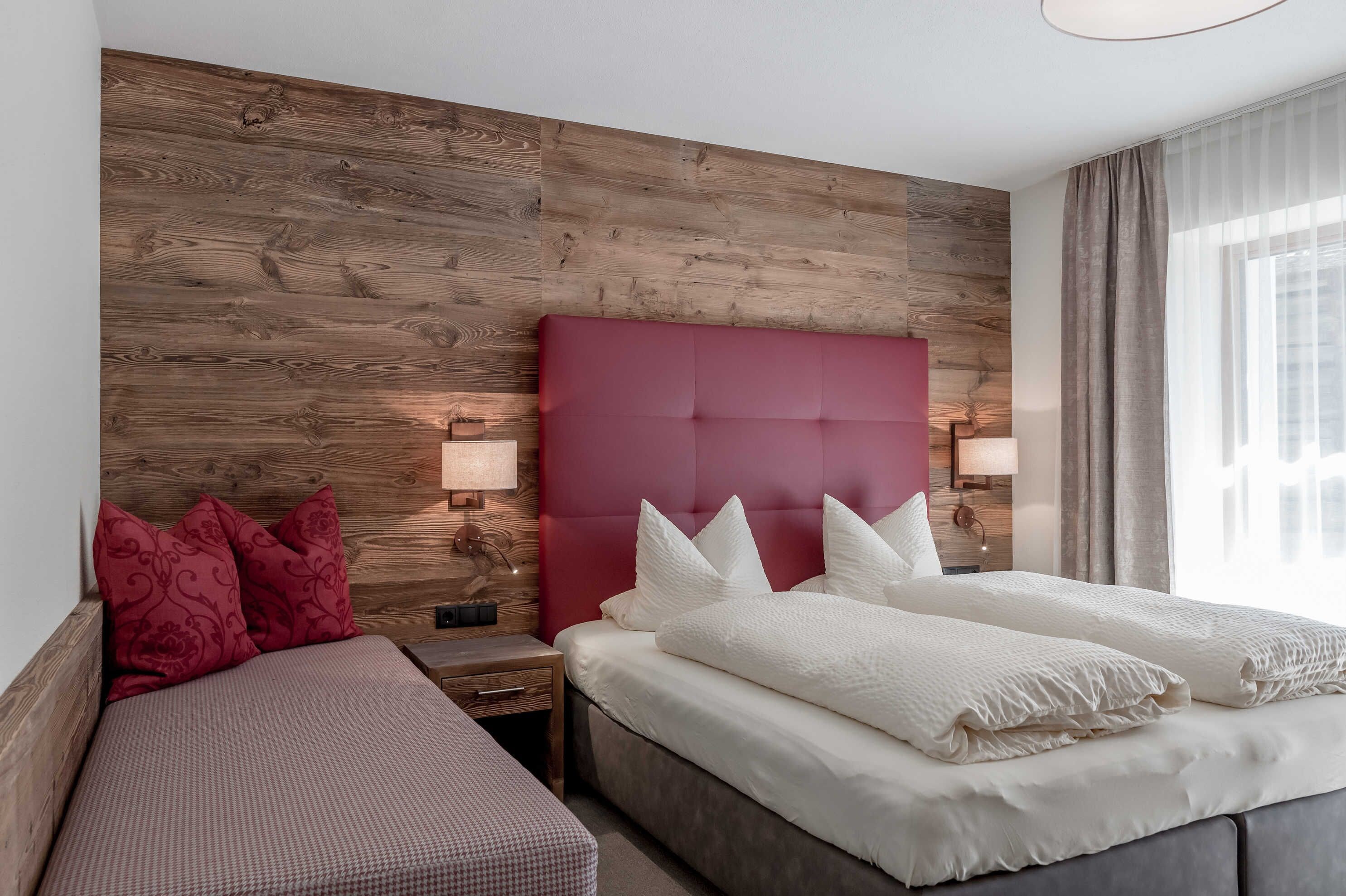 Hotelkamer met houten hoofdeinde, rode accentmuur, bed met wit beddengoed, nachtkastje met lamp en raam met gordijnen.