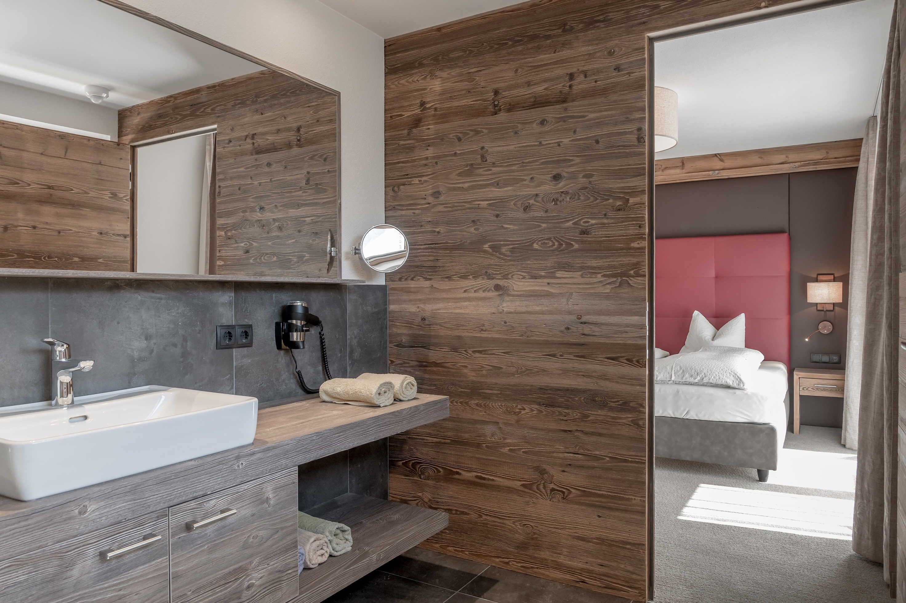 A modern bathroom with a large mirror, wooden wall paneling, a white sink, and a bed visible through an open door.
