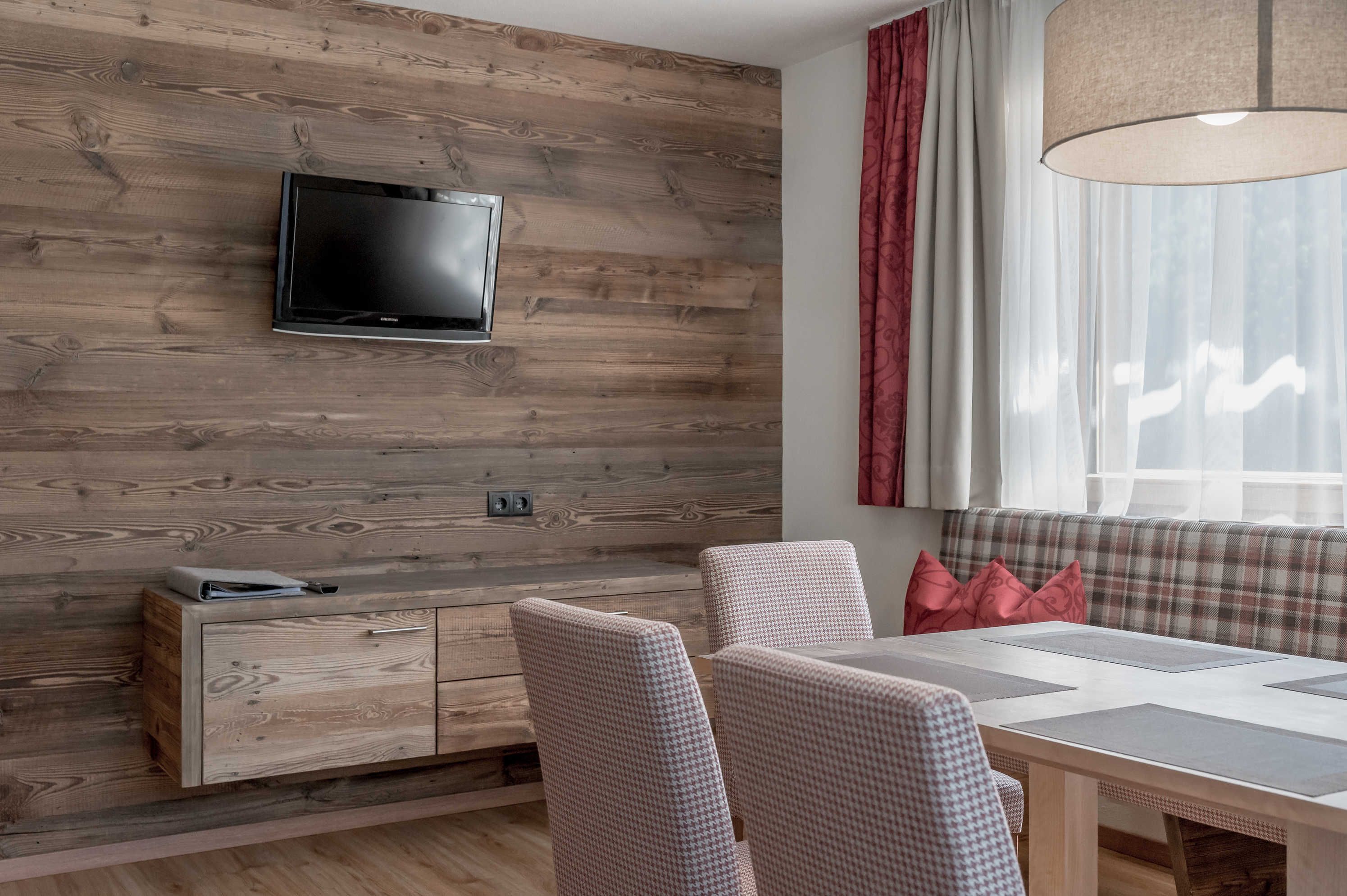 A modern living room with a wooden wall, a flat-screen TV, a dining table, and a bench with cushions.