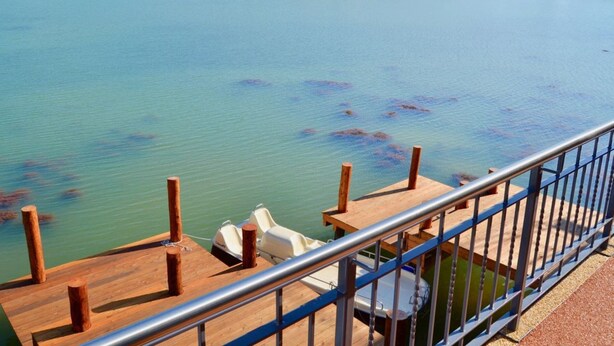 Una terraza de madera con barandilla metálica que da a un lago tranquilo con aguas cristalinas y una orilla boscosa.