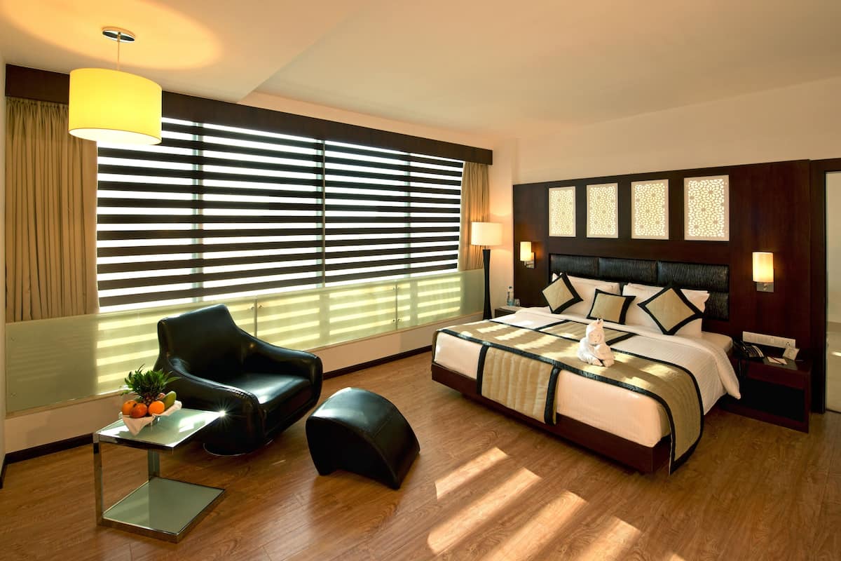 Una habitación de hotel moderna con una cama grande, un sillón de cuero negro, una mesita con frutas y un amplio ventanal con persianas.