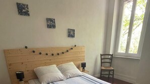 Un dormitorio con cabecera de madera, cama con ropa de cama a rayas, dos lámparas de pared, una silla y una ventana con vistas a la vegetación.