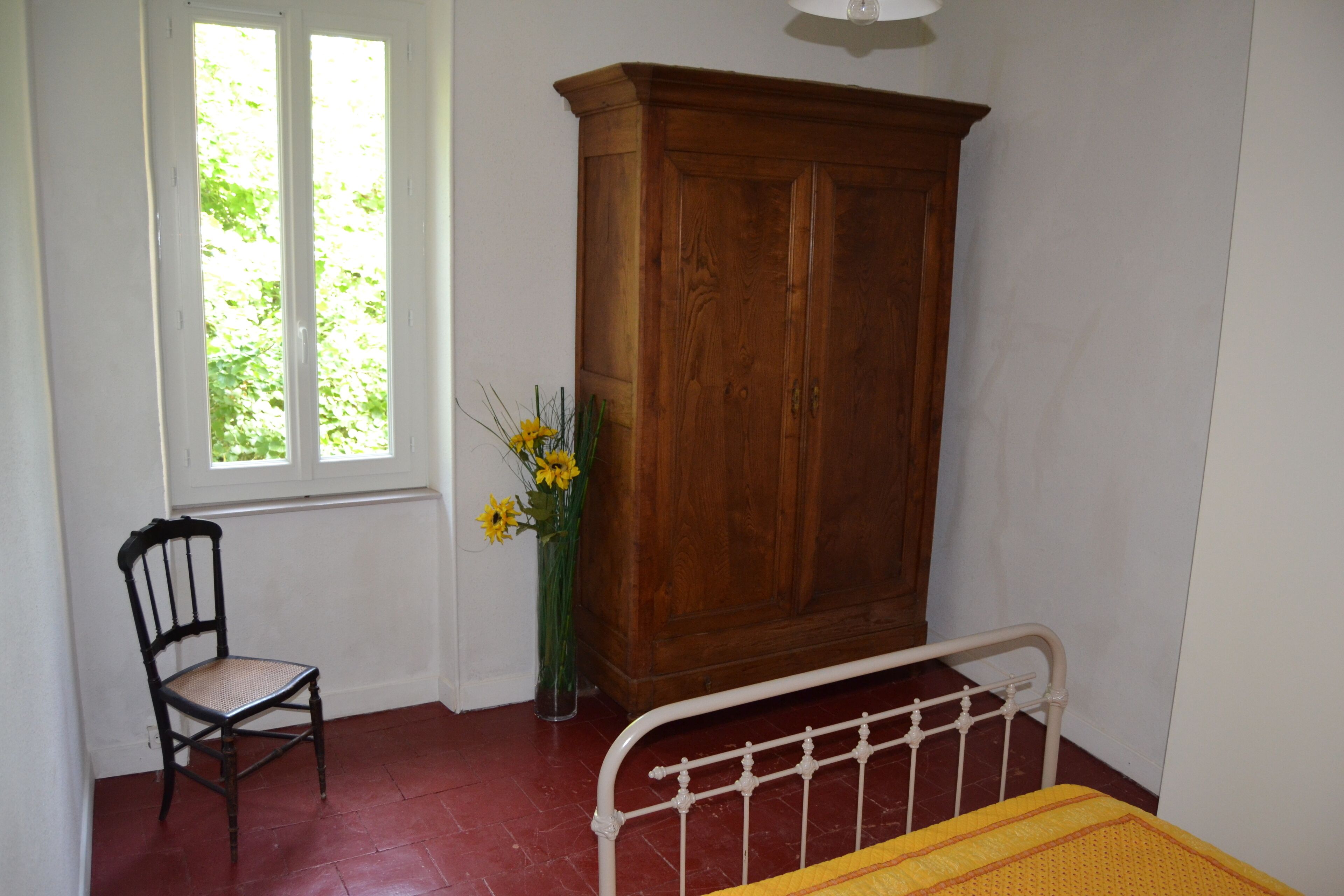 Een kamer met een houten kast, een stoel, een bed met metalen frame en een raam met uitzicht op groen.