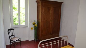 Een kamer met een houten kast, een stoel, een bed met metalen frame en een raam met uitzicht op groen.