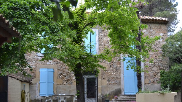 Un edificio de piedra con contraventanas azules, un árbol en frente y una pequeña mesa con sillas.
