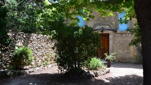 Un edificio rústico de piedra con puerta de madera y contraventanas azules, rodeado de vegetación y un camino de grava.