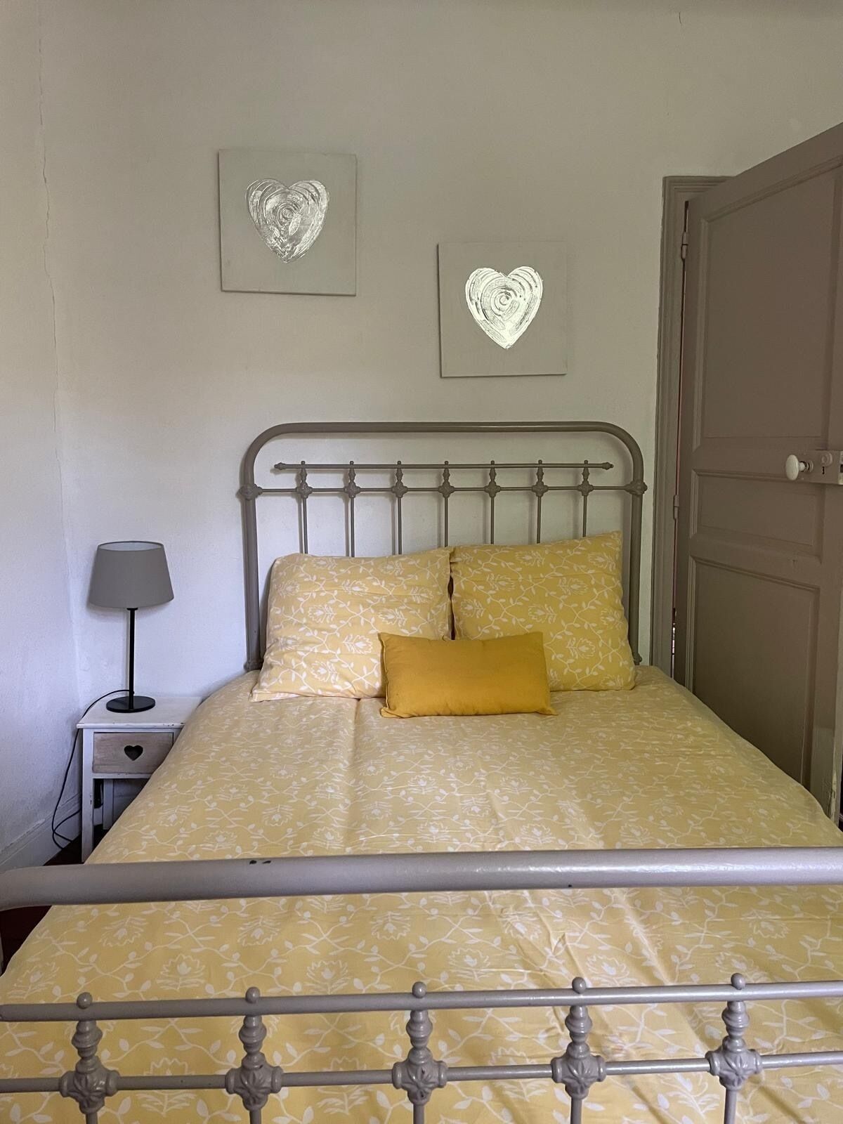 Una cama con estructura metálica, una mesita de noche con lámpara y dos adornos en forma de corazón en la pared.
