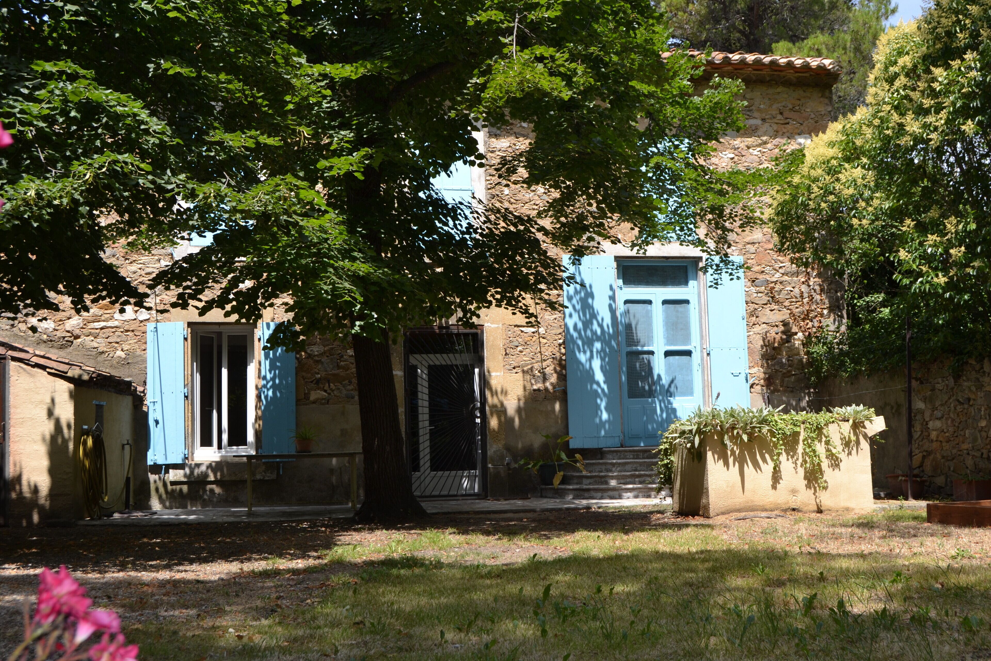 Un bâtiment rustique en pierre, doté d’une porte en bois et de volets bleus, entouré de verdure et agrémenté d’un petit jardin.