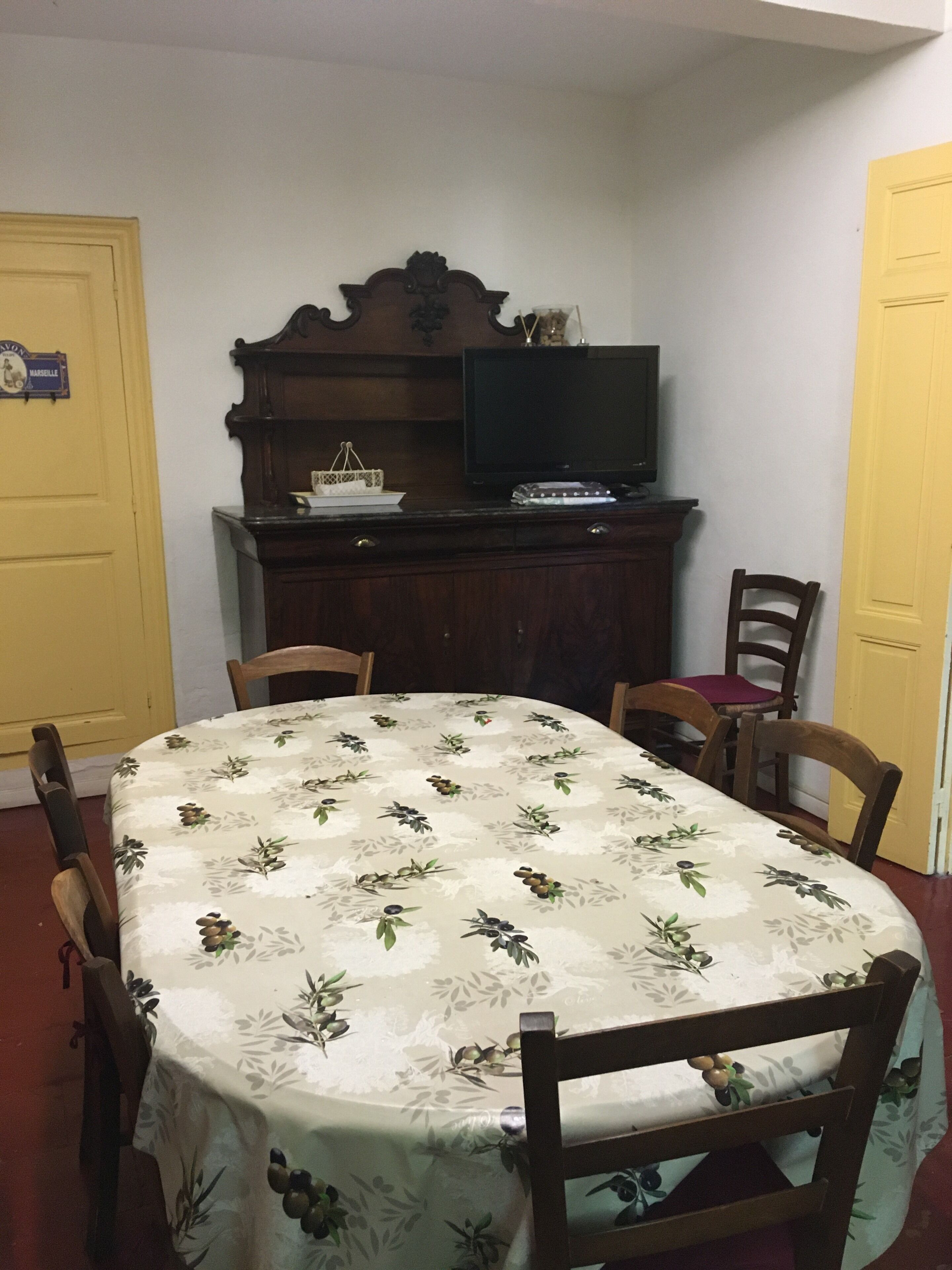Un coin repas avec une table recouverte d’une nappe à motifs floraux, entourée de chaises en bois. Un téléviseur est fixé sur un meuble en bois au-dessus de la table.