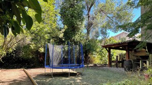 Un trampolín en un jardín con una zona de juegos de madera y una estructura de piedra.