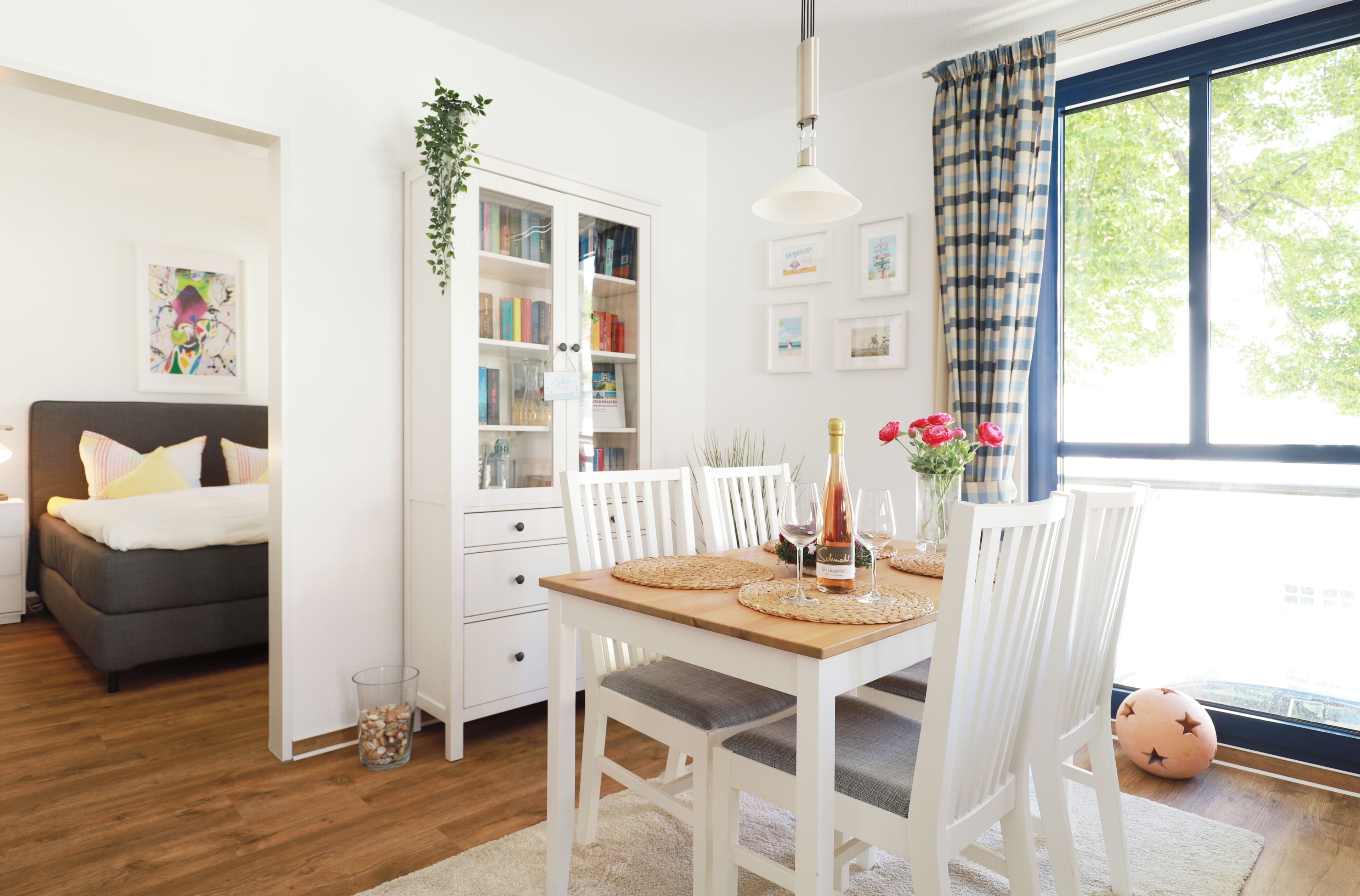 A well-lit room with a dining table set for two, a bookshelf, a sofa, and a window with curtains.