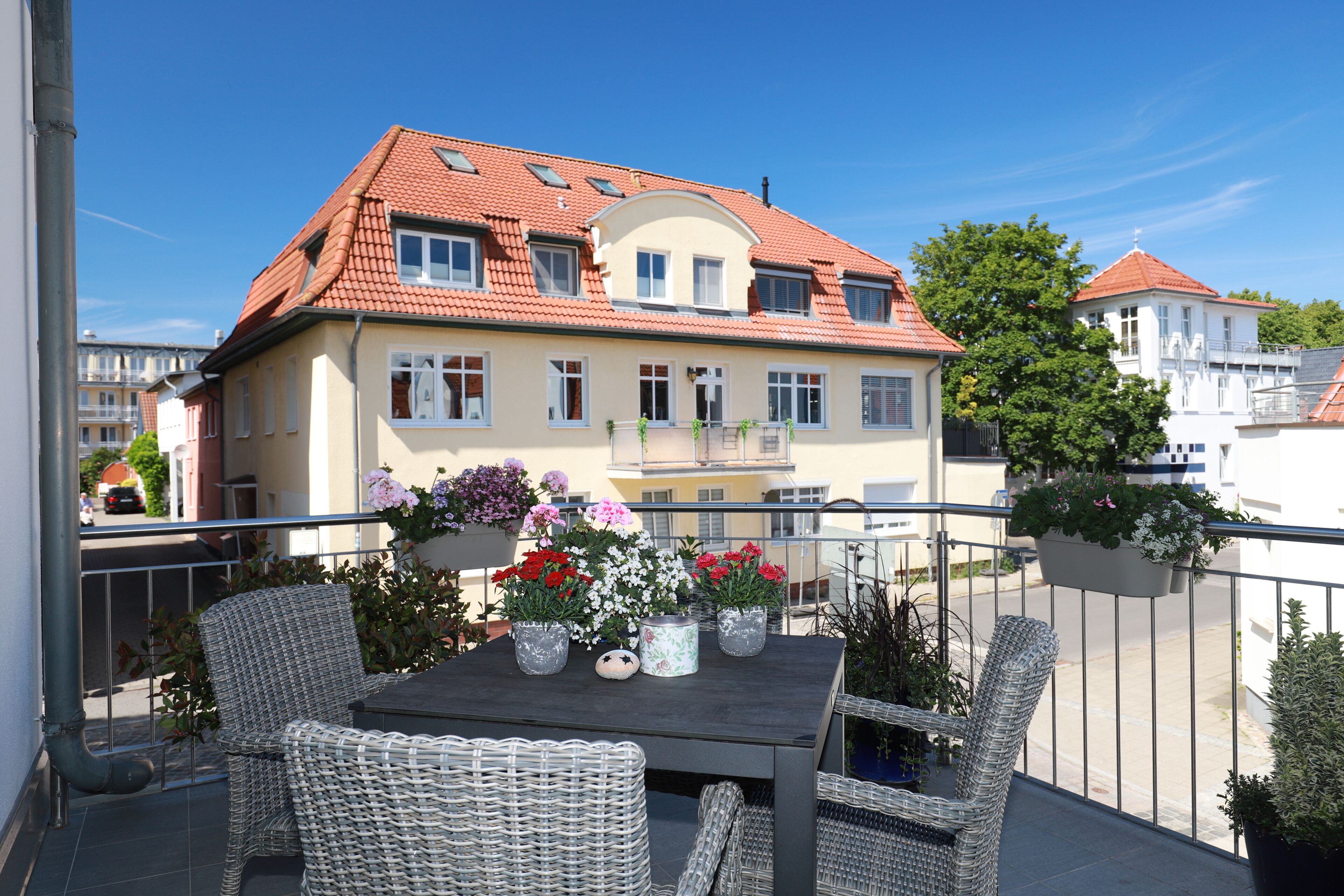 A balcony with wicker furniture, a table with potted plants, and a building with a red-tiled roof in the background.