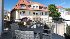 Ein Balkon mit Korbmöbeln, einem Tisch mit Topfpflanzen und ein Gebäude mit rotem Ziegeldach im Hintergrund.