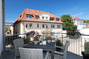 A balcony with wicker furniture, a table with potted plants, and a building with a red-tiled roof in the background.