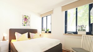 Ein Schlafzimmer mit einem Bett, Nachttischen, einem Sessel, einem Heizkörper und einem Fenster mit Blick auf Bäume.
