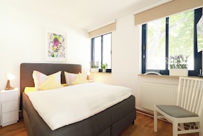 A bedroom with a bed, bedside tables, a chair, a radiator, and a window with a view of trees.