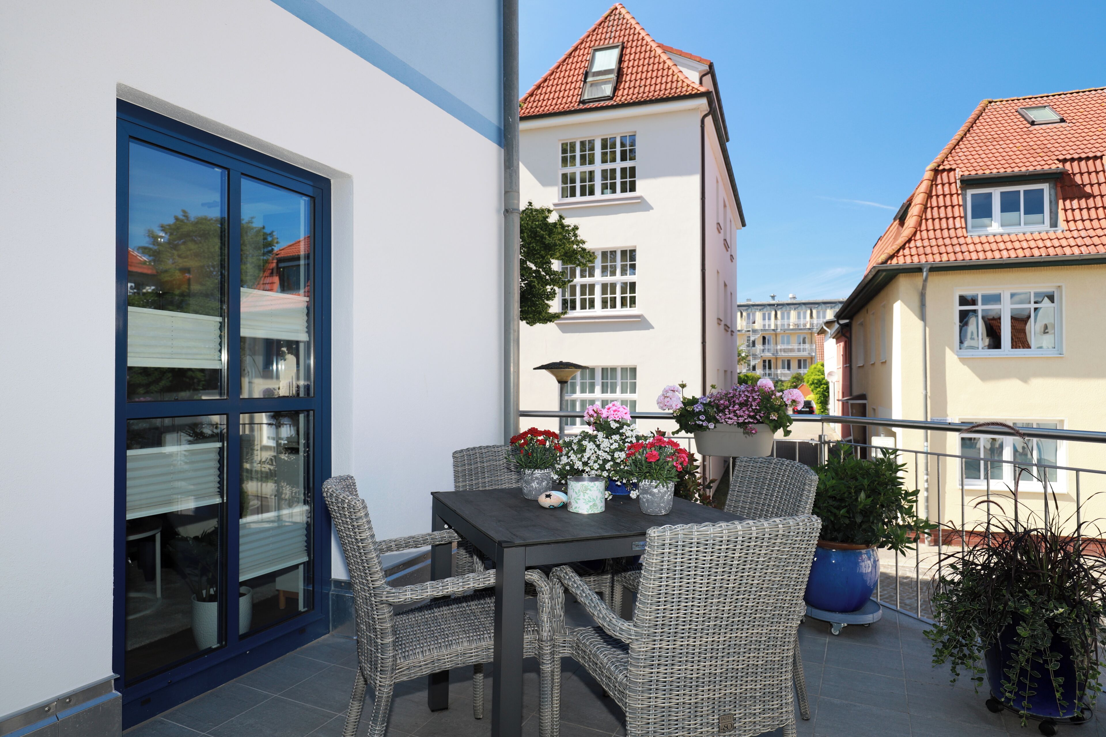 A balcony with wicker furniture, a table with flowers, and a view of neighboring buildings.