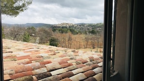 Vista dalla finestra su un tetto in tegole di terracotta, un pendio con alberi sparsi e un villaggio in lontananza.