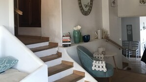 Una scala conduce a un secondo piano, una poltrona blu con copriletto in pizzo, uno specchio appeso alla parete e un tavolino con libri e oggetti decorativi.