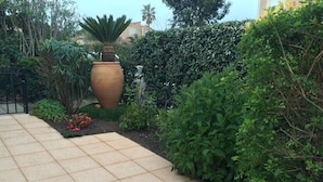 Un chemin pavé mène à un grand pot en terre cuite, entouré d’une végétation luxuriante et d’un petit parterre de fleurs.