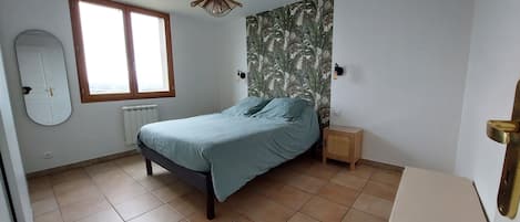 A bedroom with a bed, a window, a radiator, a small bedside table, and a wall-mounted light fixture.