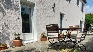 Un bâtiment à l’extérieur blanc, avec une porte et des fenêtres. On y trouve une table et des chaises à l’extérieur.