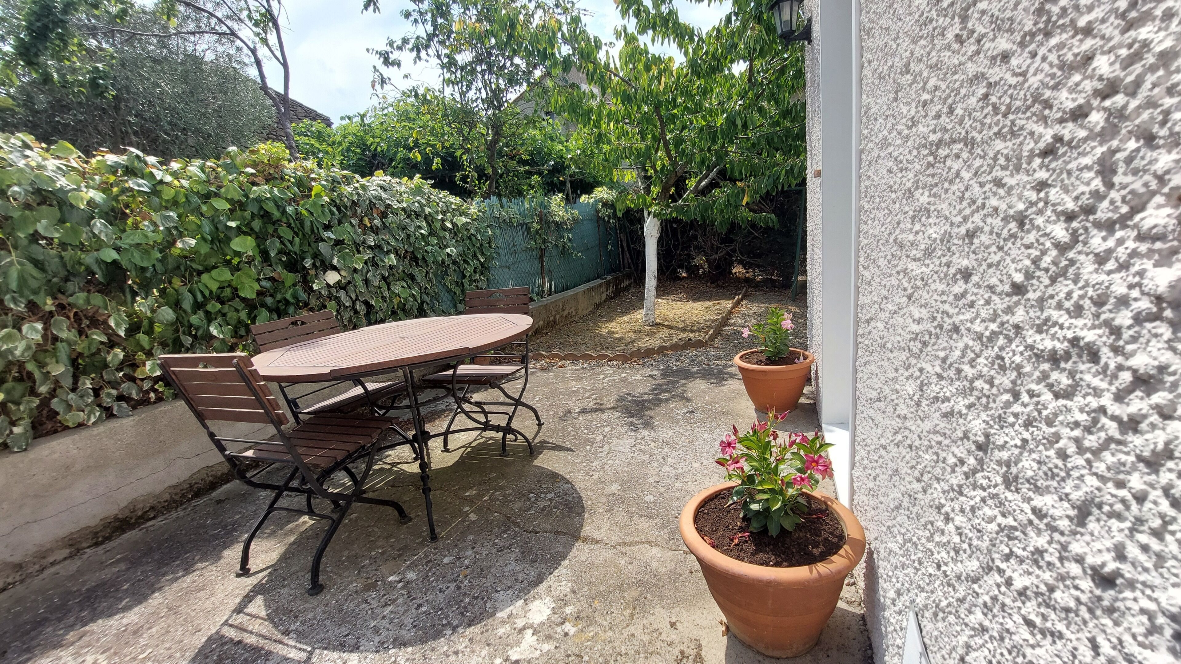 An outdoor patio with a wooden table and chairs, surrounded by potted plants and a hedge.
