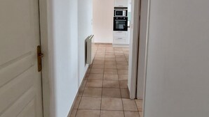 Un couloir aux murs blancs, doté d’un sol carrelé, et où l’on aperçoit une cuisine au fond.