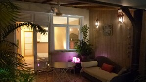 A cozy patio area with a round table and chairs, a cushioned seating area, and a wooden wall with a window. - Studio in Didier FORT DE FRANCE (Fort de France)