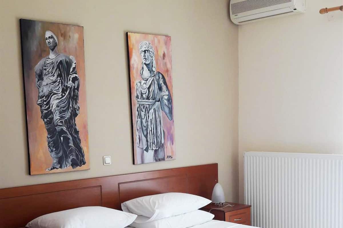 A hotel room with a wooden bed, white linens, two paintings on the wall, a bedside table, and a radiator.