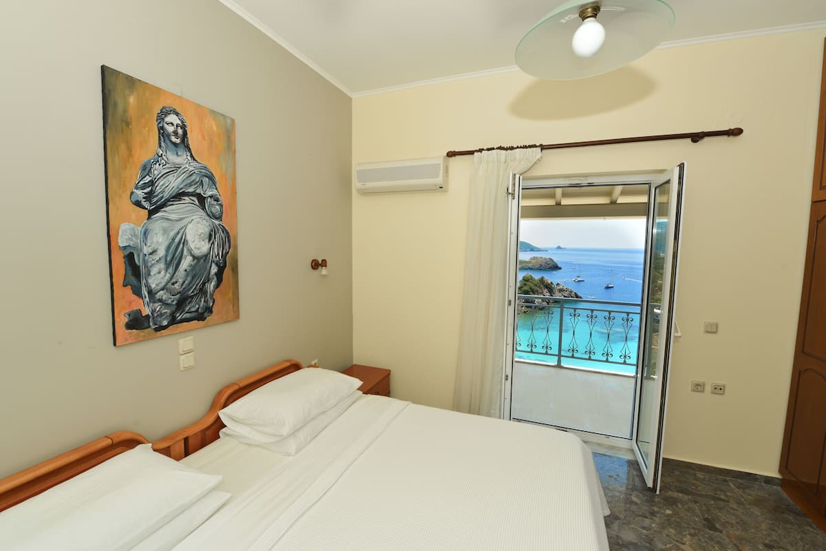 A hotel room with a bed, a painting, an air conditioning unit, and a balcony with a view of the sea.