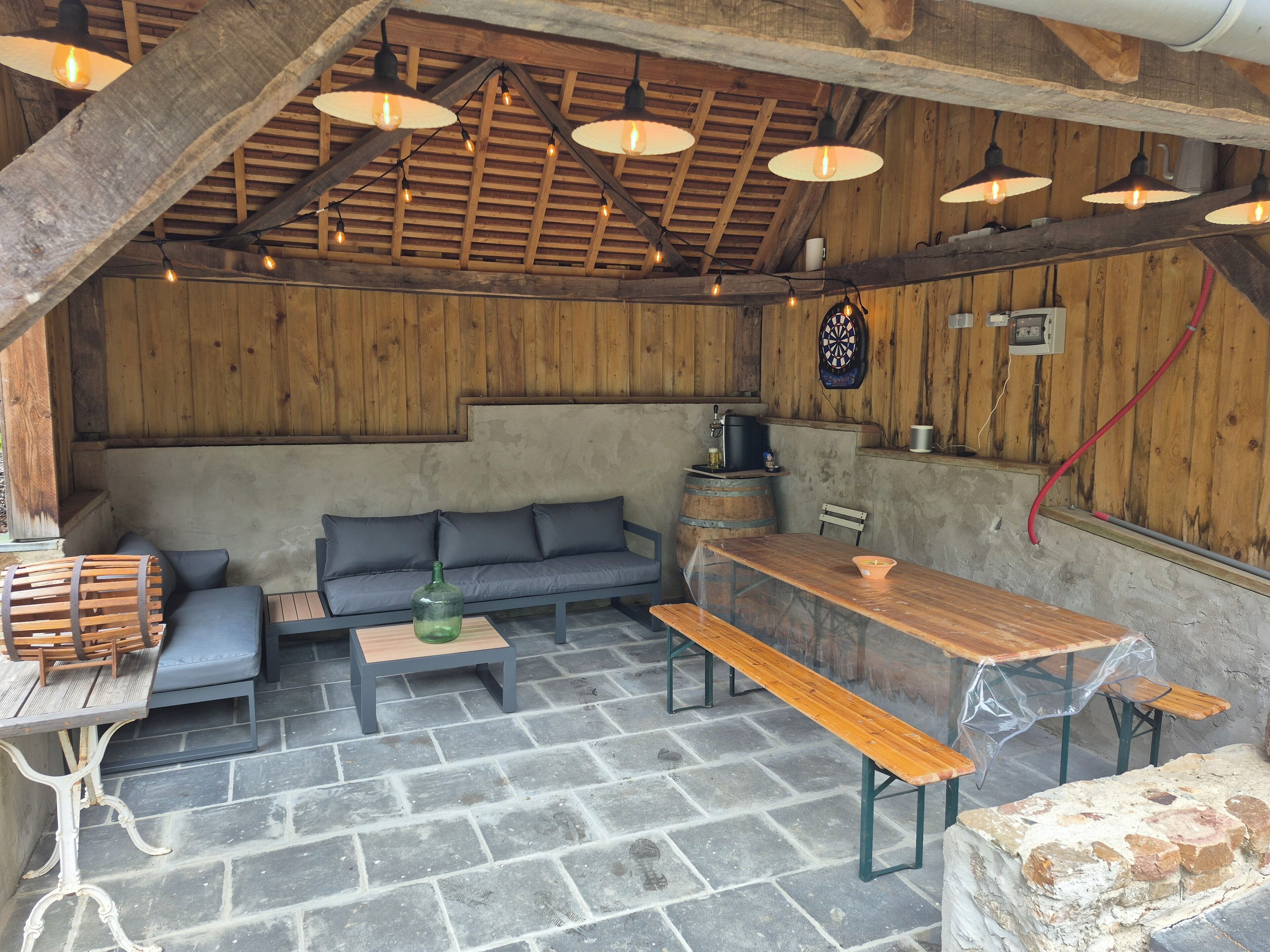 A rustic interior with a stone floor, wooden beams, a sofa, a table, and a dartboard.