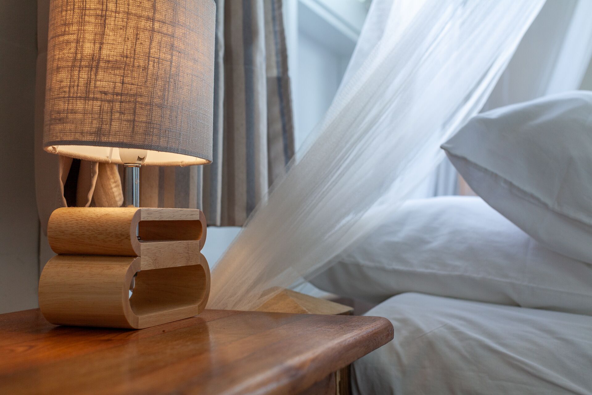 Ein Nachttisch aus Holz mit einer Lampe, ein Bett mit weißer Bettwäsche und ein durchsichtiger weißer Vorhang.
