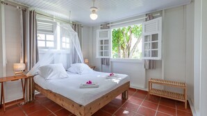 A bedroom with a wooden bed, white curtains, a bedside table with a lamp, a window with shutters, and a bench. - Villa Vue Sauvage in Tartane, in the Caravelle nature reserve. (LA TRINITÉ)
