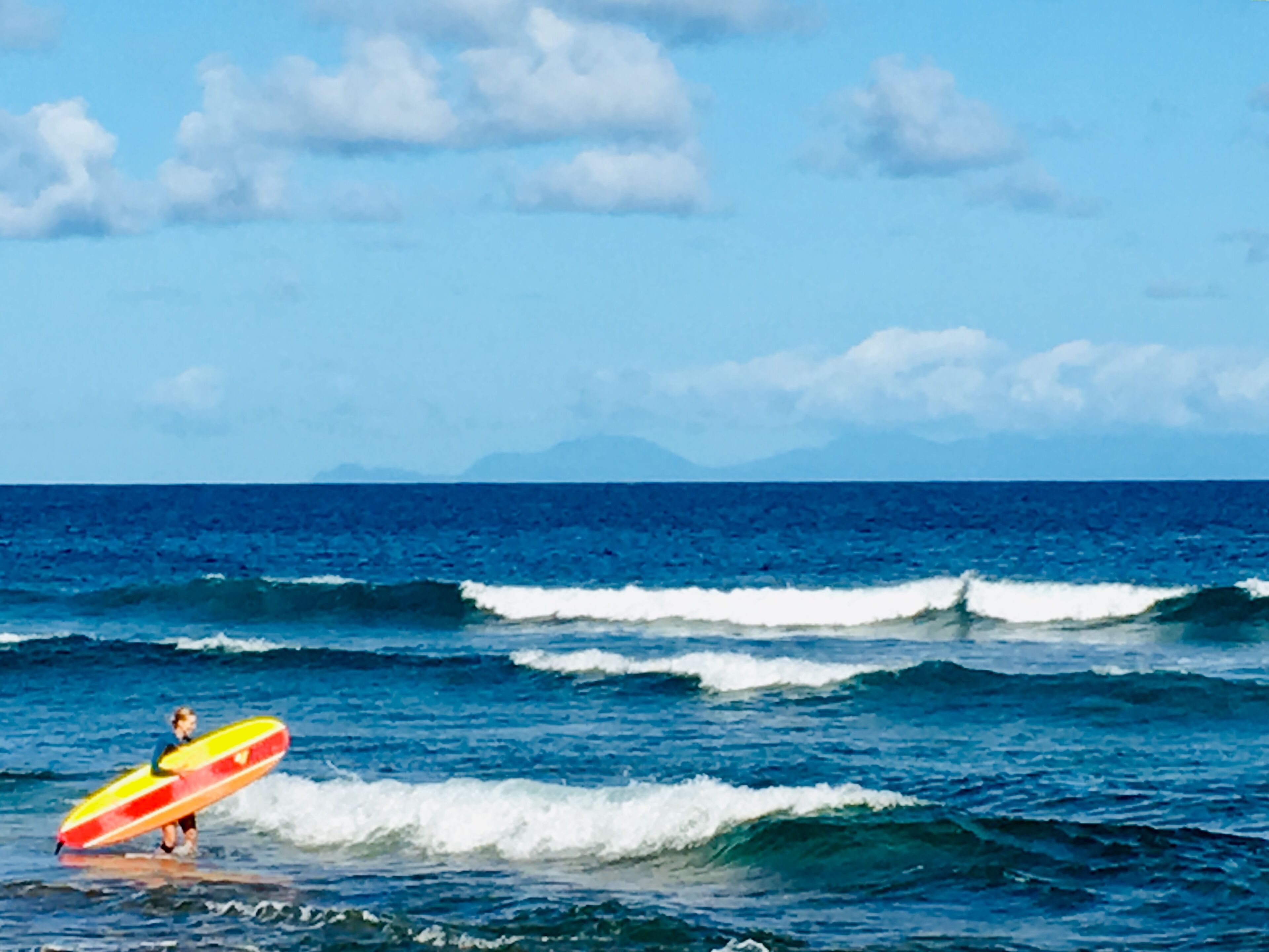 Une personne tenant une planche de surf dans l’océan, avec des vagues et un ciel dégagé.