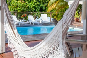 Uma rede com vista para uma piscina, com cadeiras de descanso brancas e vegetação ao fundo.