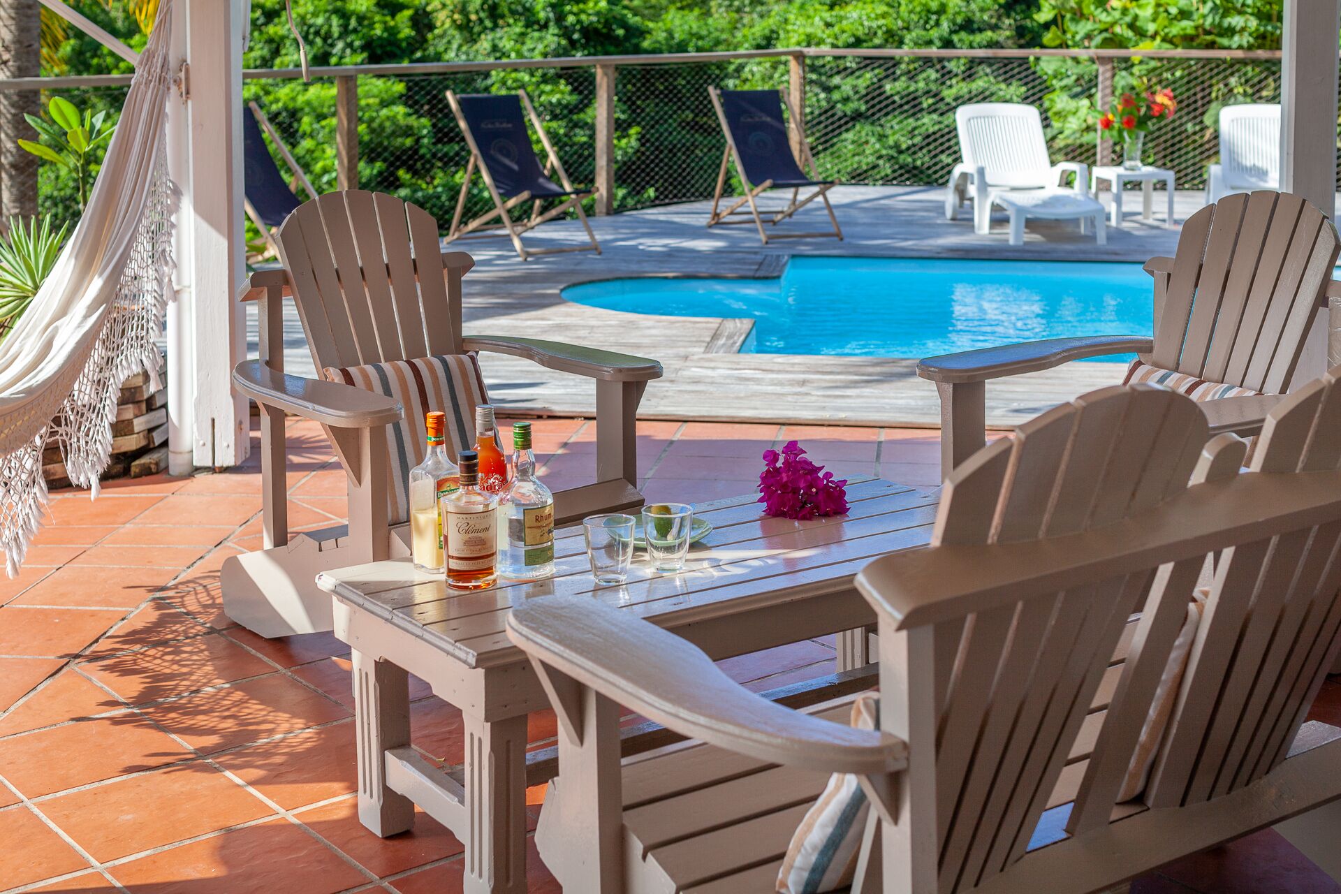 A poolside area with wooden chairs, a table with bottles and glasses, and a hammock.