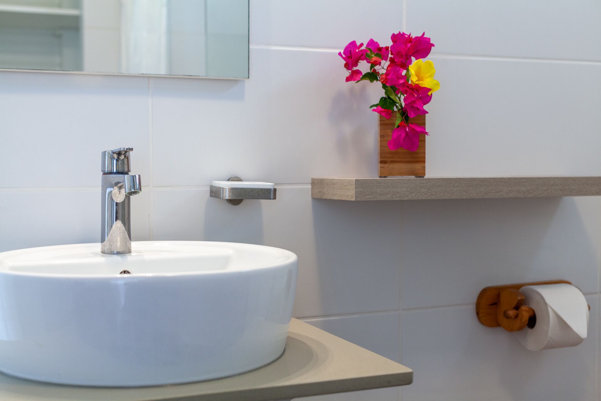 Un baño con un lavamanos blanco, un espejo y un estante de madera con flores.
