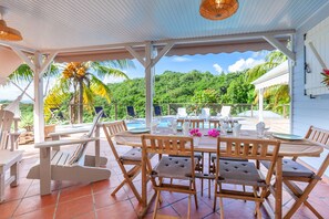 Uma área de refeições ao ar livre coberta, com mobiliário de madeira e piscina ao fundo.