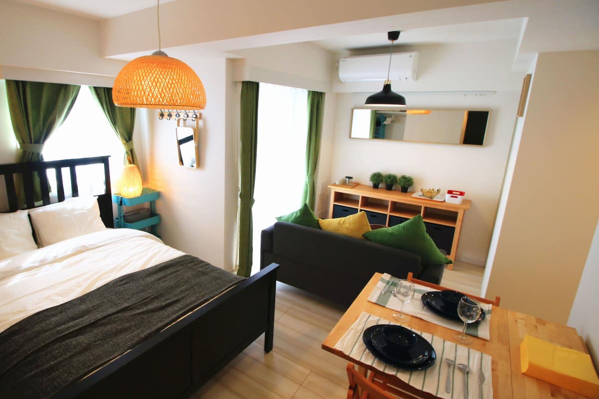 Quarto com cama, mesa de cabeceira, luminária e vista para uma sala de estar com sofá e mesa de jantar posta para dois.